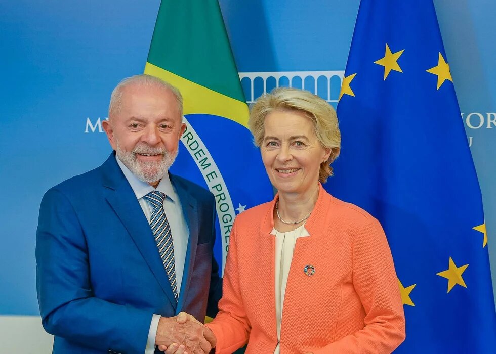 Foto: Luiz Inácio Lula da Silva und Ursula von der Leyen geben sich die Hand. Im Hintergrund die Flaggen Brasiliens und der EU.